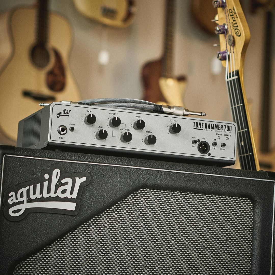 Angled front view of Aguilar Tone Hammer 700 Bass Amp Head on top of speaker cab with bass leaning on it and guitars in the background