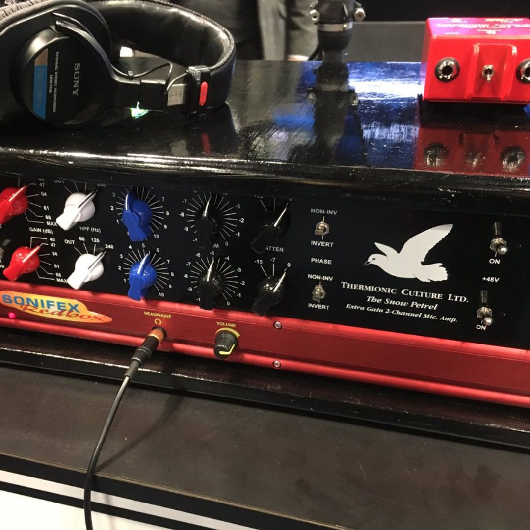 Close up of black rackmount effect on top of red microphone preamplifier on top of desk with headphones and red DI box