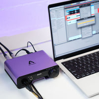 Purple audio interface device connected to a laptop with music software open on a white surface.