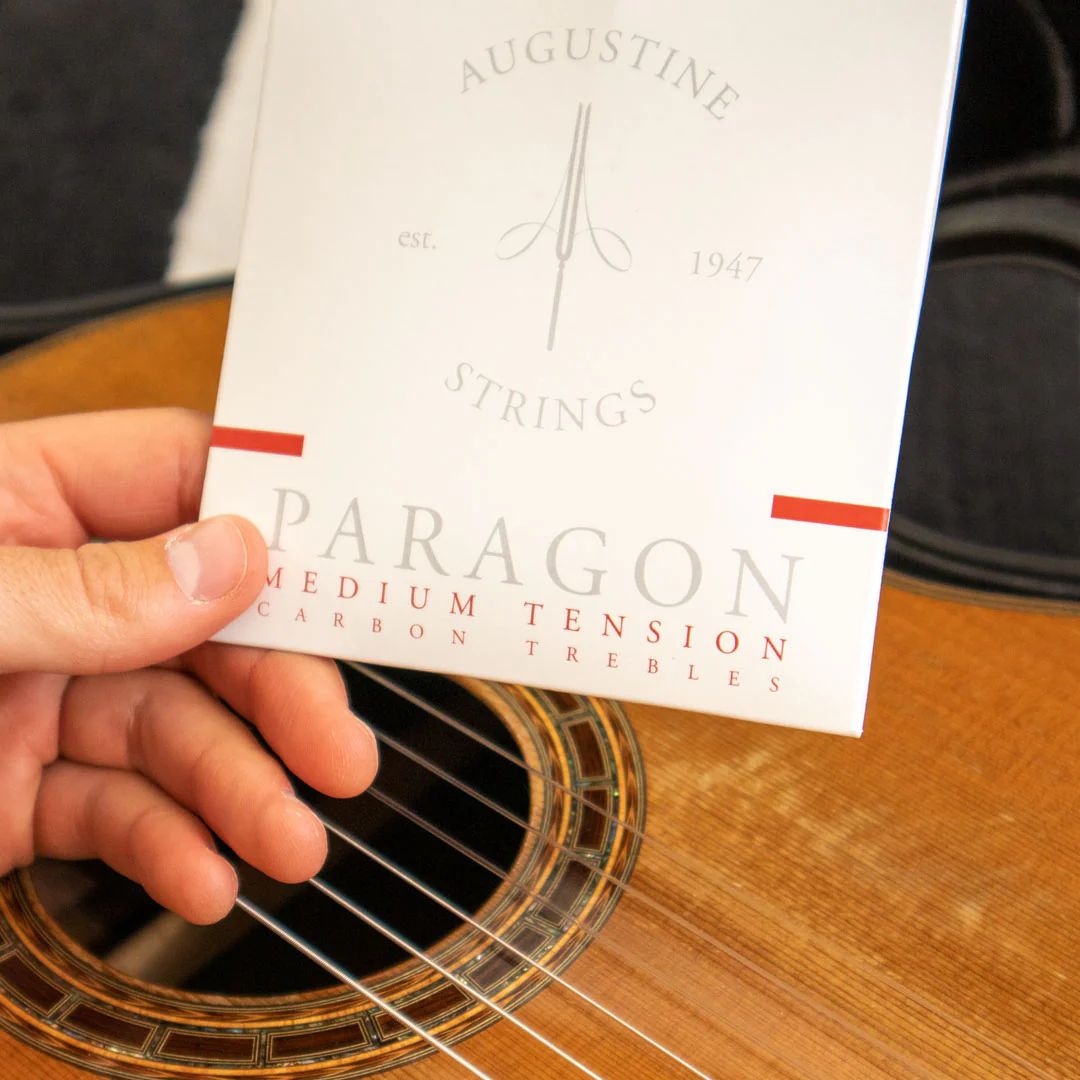 Hand holding a card labeled 'Augustine Strings Paragon Medium Tension Trebles' in front of an acoustic guitar.