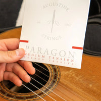 Hand holding a card labeled 'Augustine Strings Paragon Medium Tension Trebles' in front of an acoustic guitar.