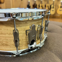 Snare drum with wooden finish and chrome hardware on a brown surface, with guitars in the background.