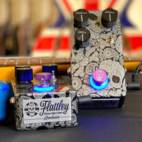 Front and top view of pair of Flattley Revolution Dynamic Overdrive Pedals on black surface with guitars in background