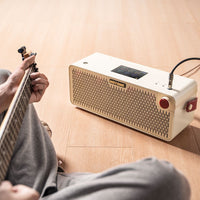 Person playing guitar with a white and beige portable speaker on a wooden floor