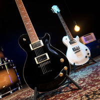 Black electric guitar on a stand with another guitar and drum set in the background.
