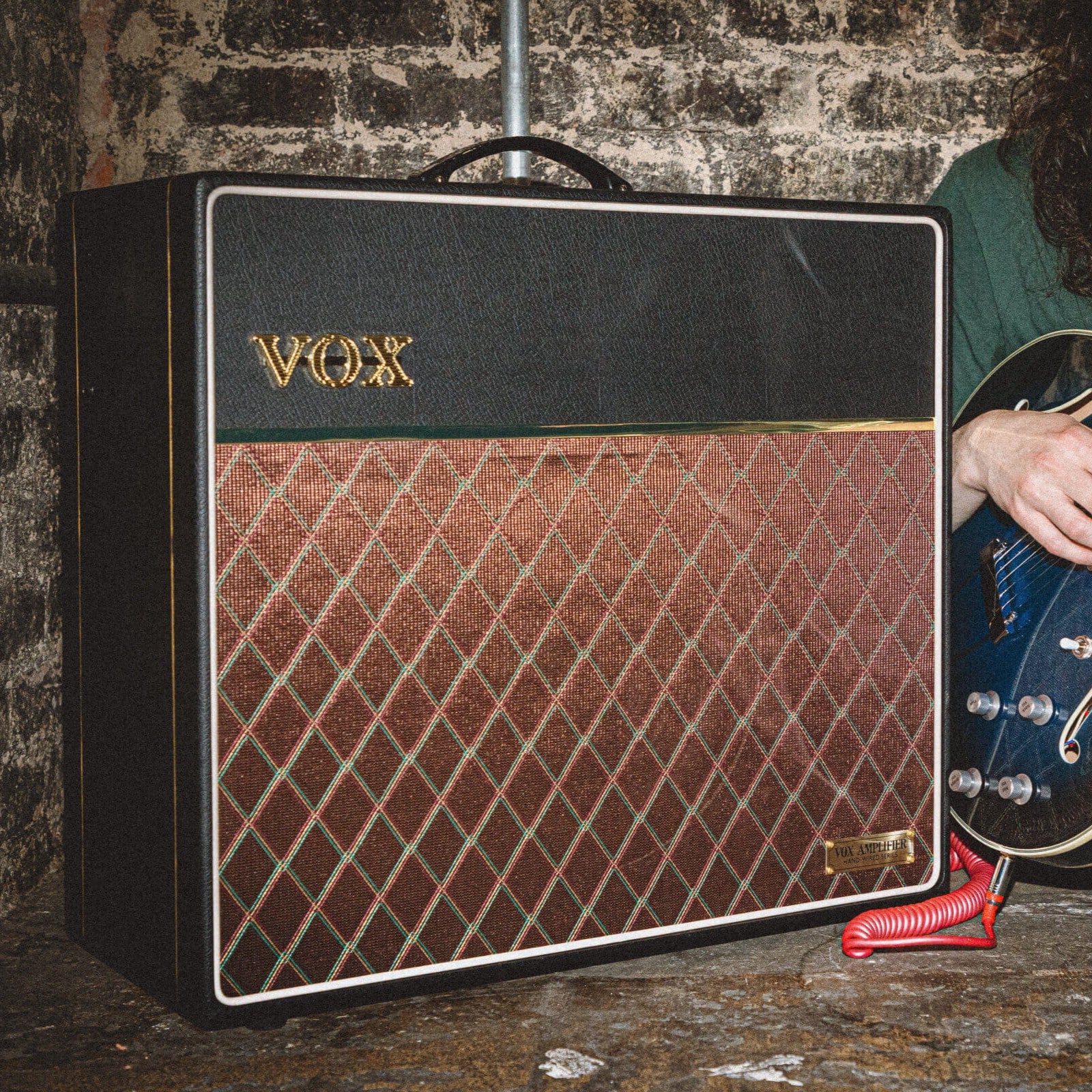 Angled front view of Vox AC15 Hand-Wired Greenback Guitar Amplifier with guitarist beside it and a brickwall background